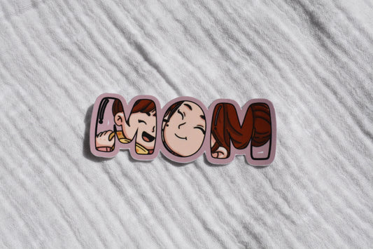 A die-cut sticker spelling 'MOM' in stylized cartoon letters, placed on a textured white fabric background.