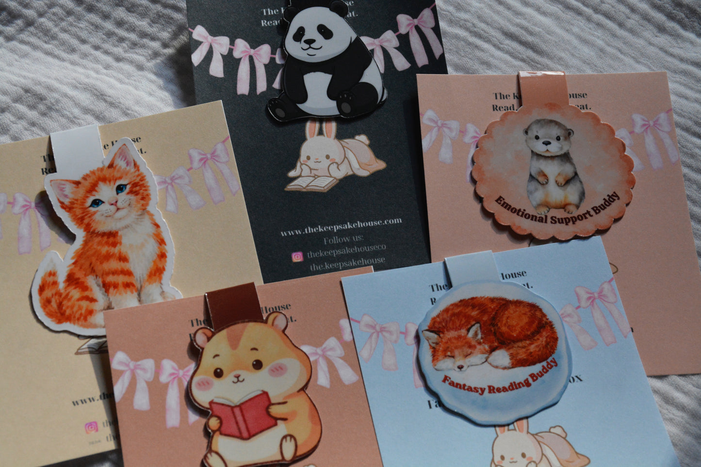A collection of cute animal-themed magnetic bookmarks displayed on their custom backing cards. The cards feature pastel colors with pink bows and include designs of an orange tabby kitten, a black-and-white panda, a sleeping hamster reading a book, a puppy labeled 'Emotional Support Puppy', and a sleeping fox labeled 'Fantasy Reading Buddy'.