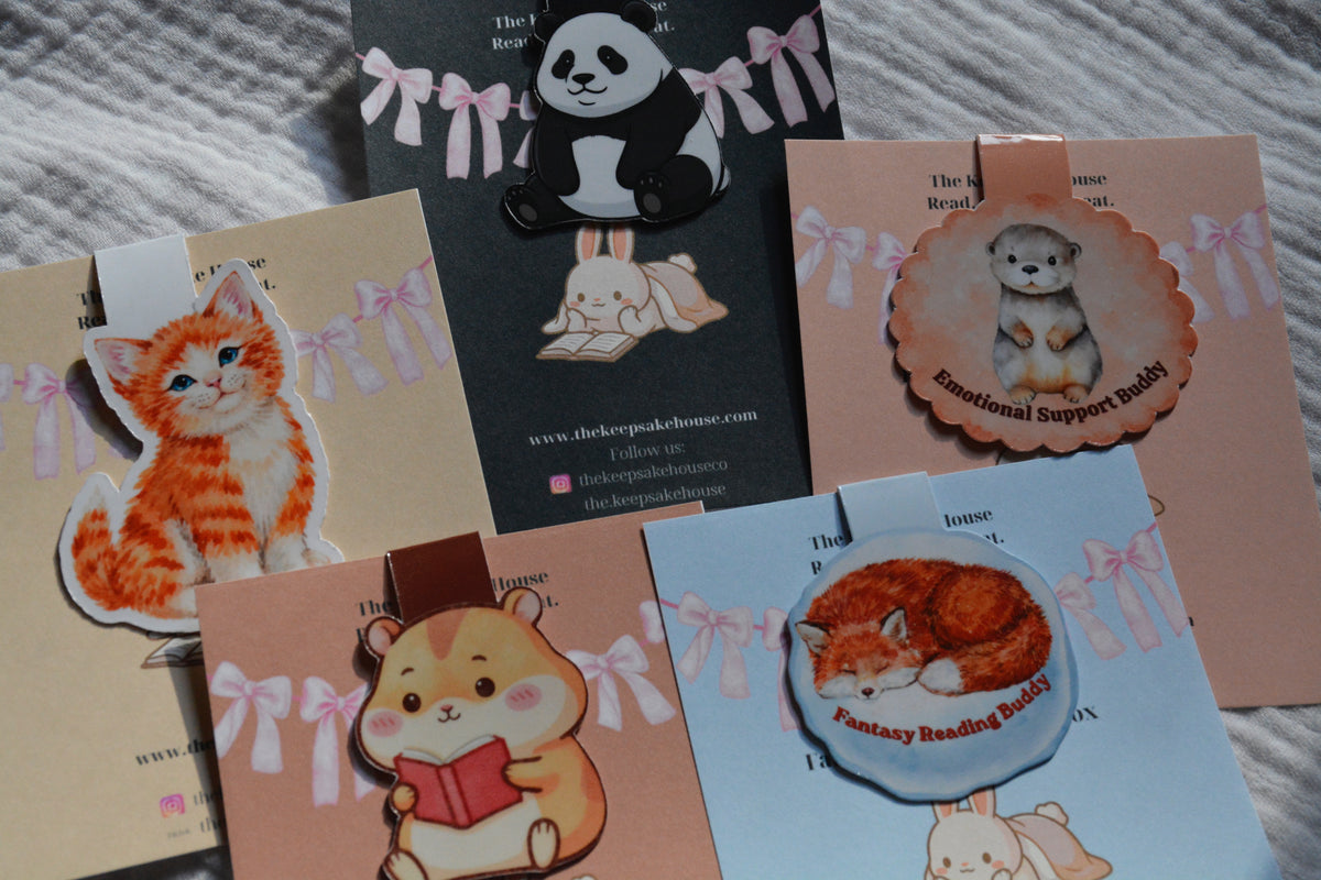 A collection of cute animal-themed magnetic bookmarks displayed on their custom backing cards. The cards feature pastel colors with pink bows and include designs of an orange tabby kitten, a black-and-white panda, a sleeping hamster reading a book, a puppy labeled 'Emotional Support Puppy', and a sleeping fox labeled 'Fantasy Reading Buddy'.