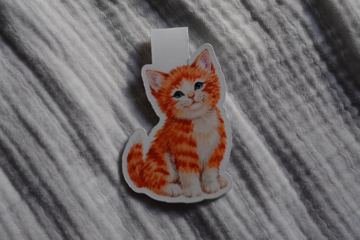 A die-cut magnetic bookmark featuring an orange and white kitten illustration, placed on a textured gray fabric background.