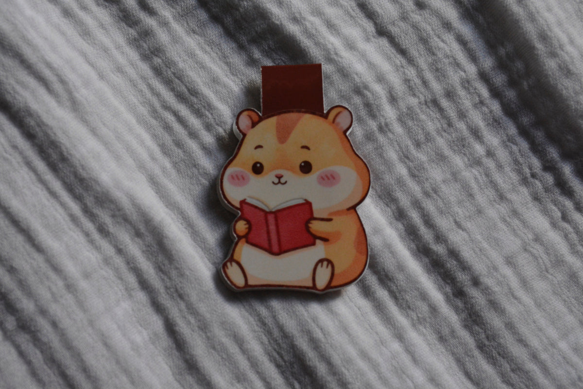 A small cartoon hamster magnetic bookmark holding a red book, photographed on a textured gray fabric background.