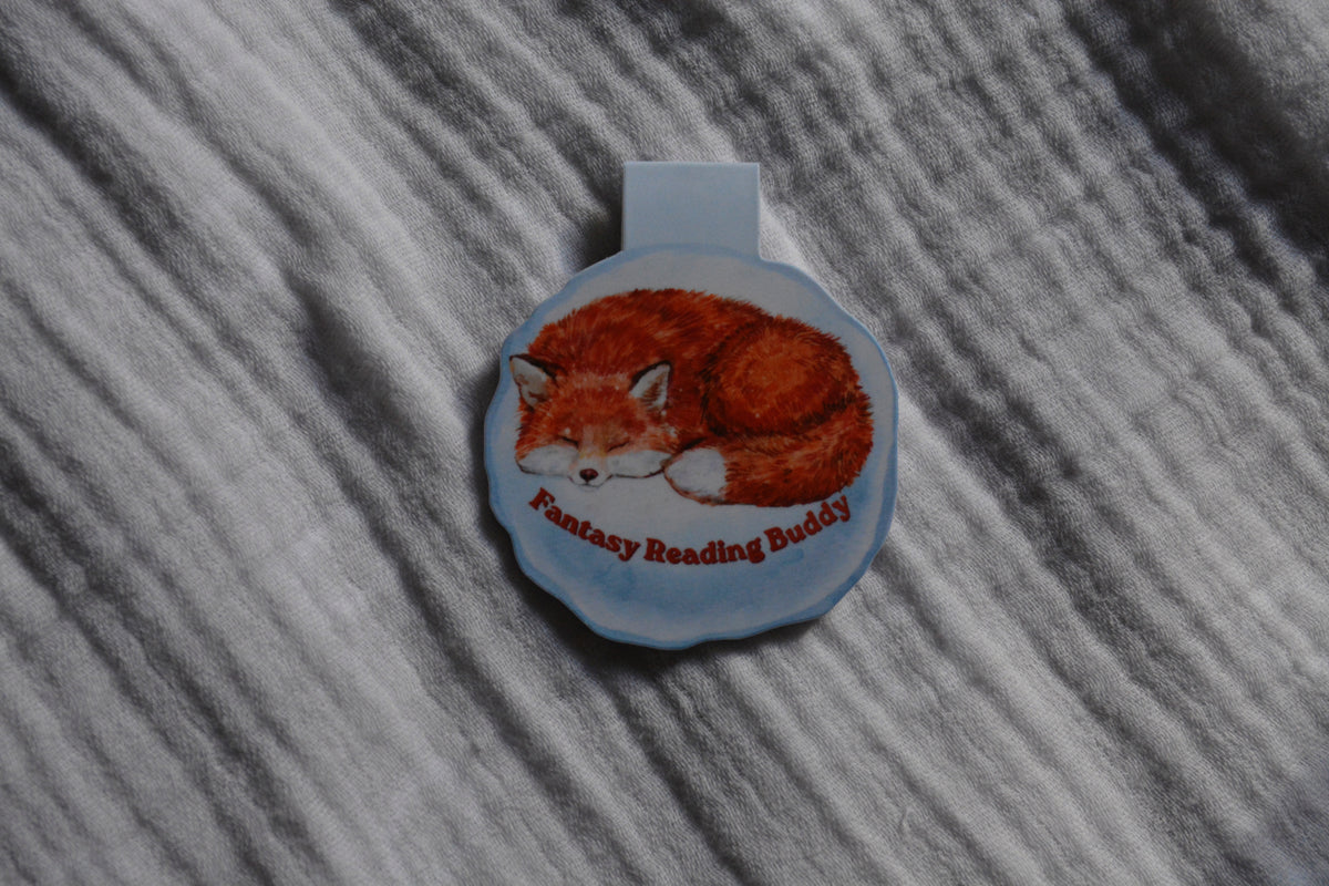 A round magnetic bookmark featuring an illustration of a sleeping orange fox and the text 'Fantasy Reading Buddy' on a textured gray fabric background.