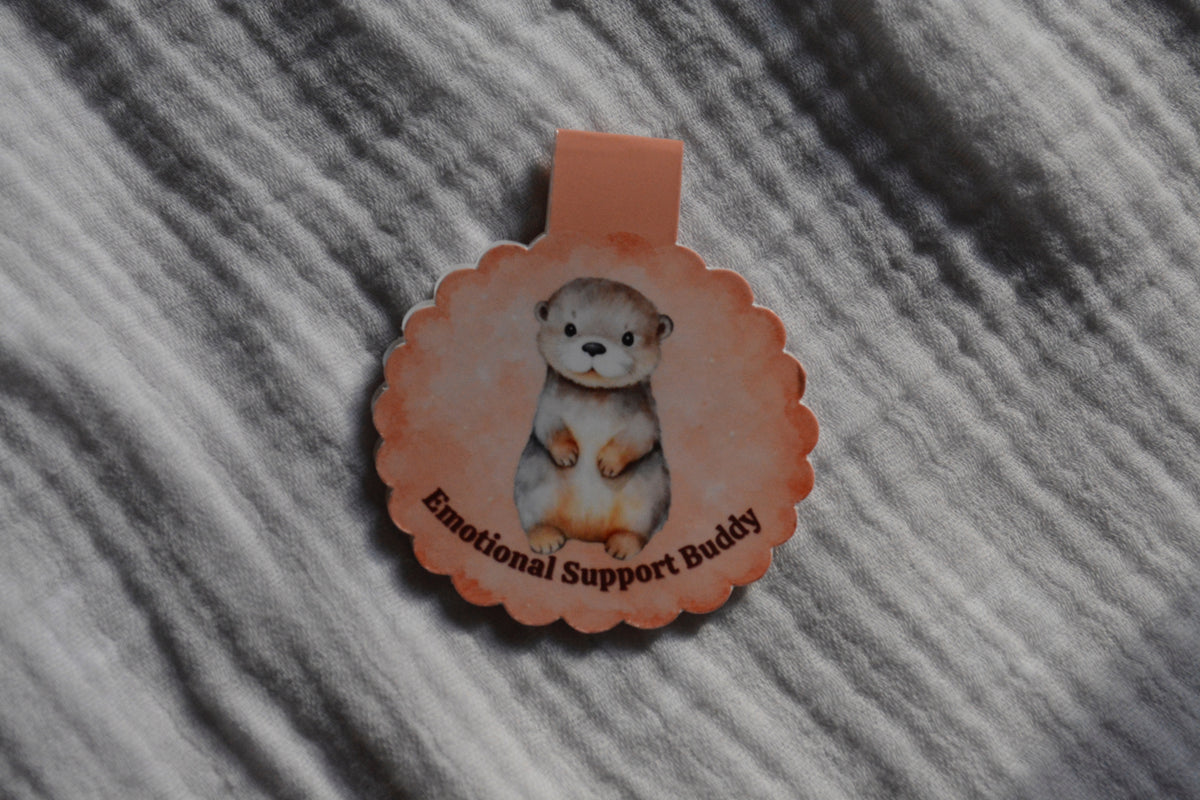 A scalloped circular magnetic bookmark cartoon otter illustration and the text 'Emotional Support Buddy' on a textured gray fabric background.