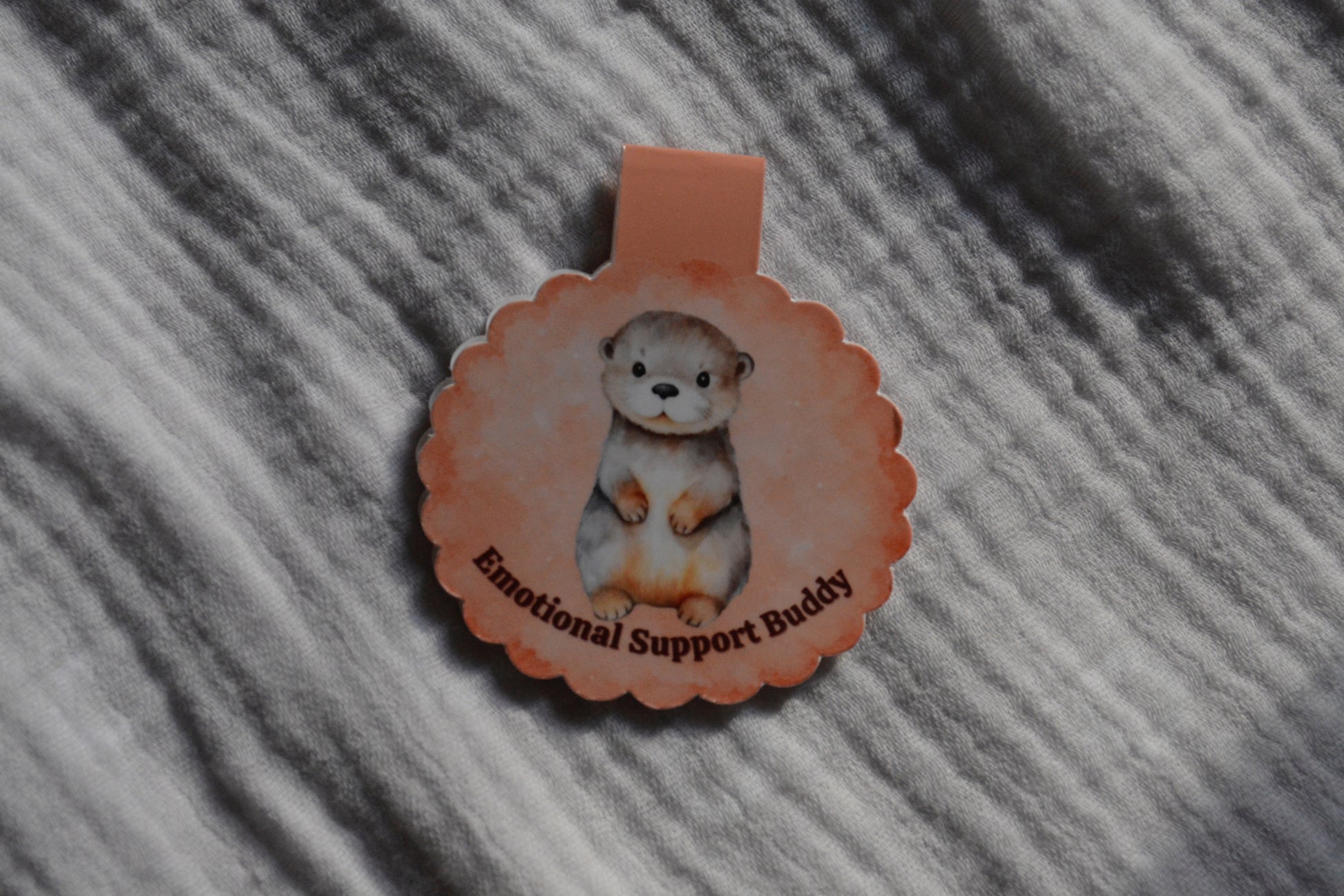 A scalloped circular magnetic bookmark cartoon otter illustration and the text 'Emotional Support Buddy' on a textured gray fabric background.