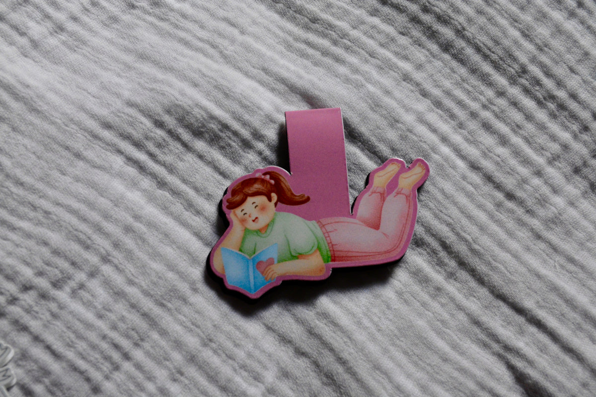  a girl reading a book magnetic bookmark featuring a pink background with a book, placed on a gray textured fabric surface.