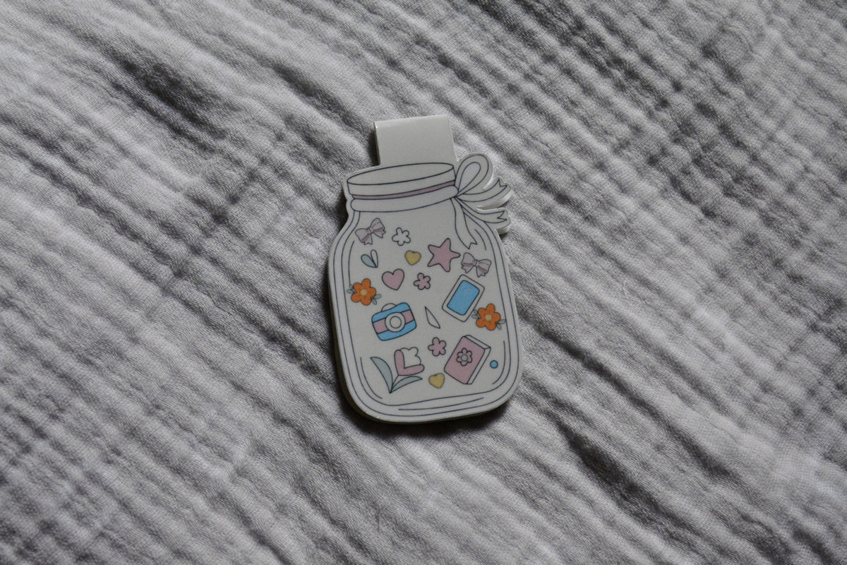 A magnetic bookmark shaped like a mason jar, decorated with pastel-colored doodles including stars, hearts, flowers, and small objects, placed on a gray textured fabric background.