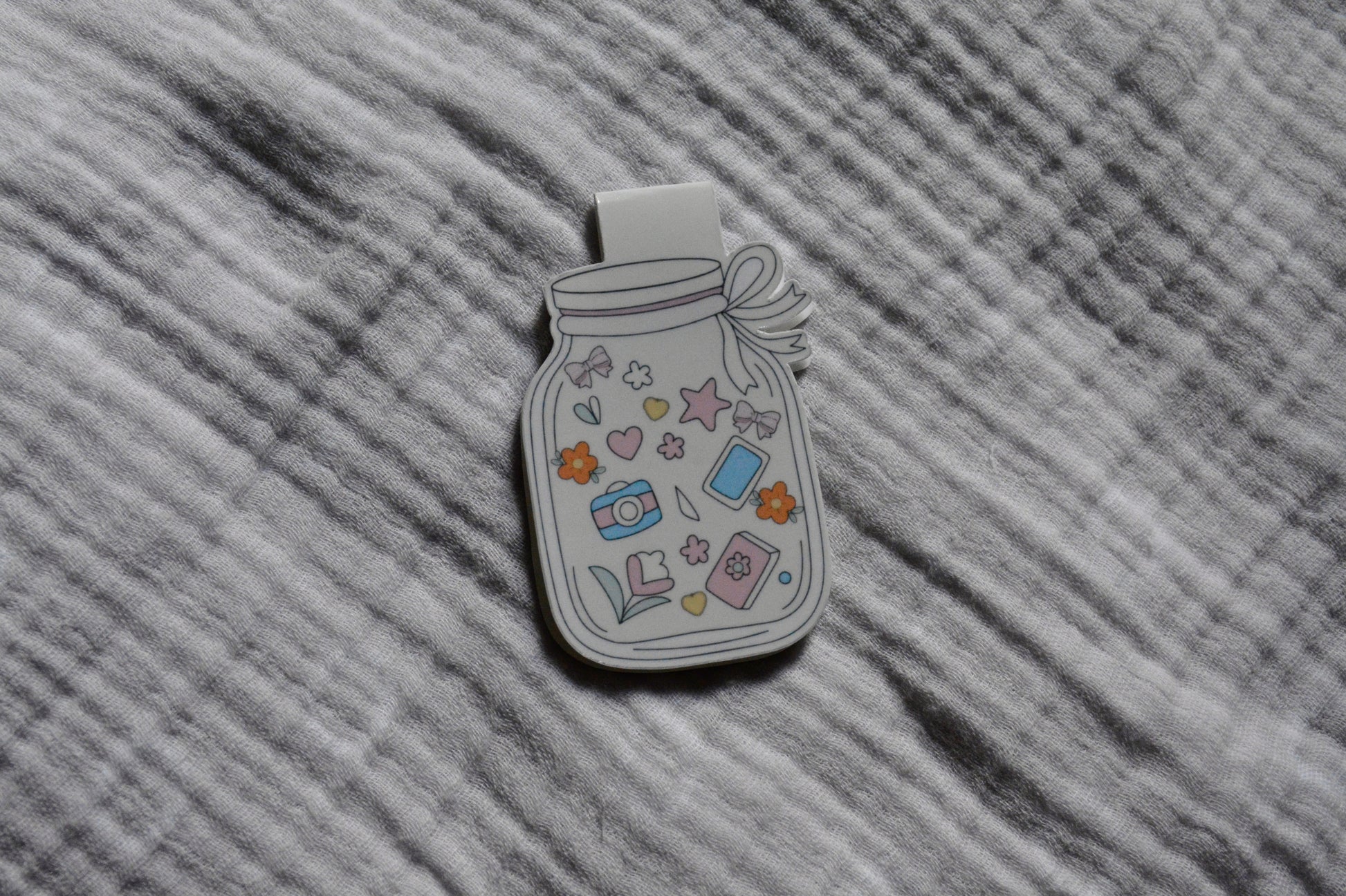 A magnetic bookmark shaped like a mason jar, decorated with pastel-colored doodles including stars, hearts, flowers, and small objects, placed on a gray textured fabric background.