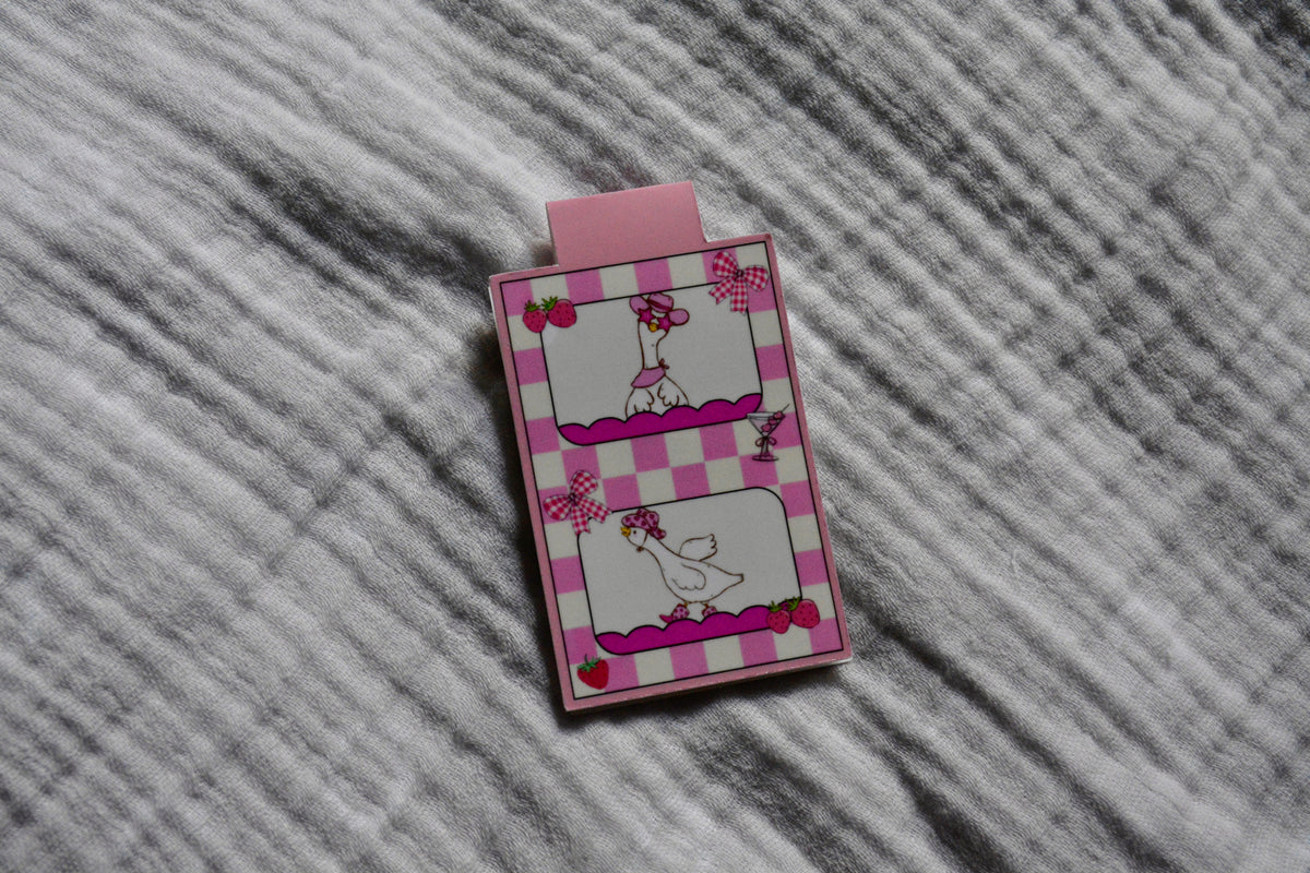 A small pink and white illustrated magnetic bookmark with cartoon silly goose illustrations, placed on a gray textured fabric background.