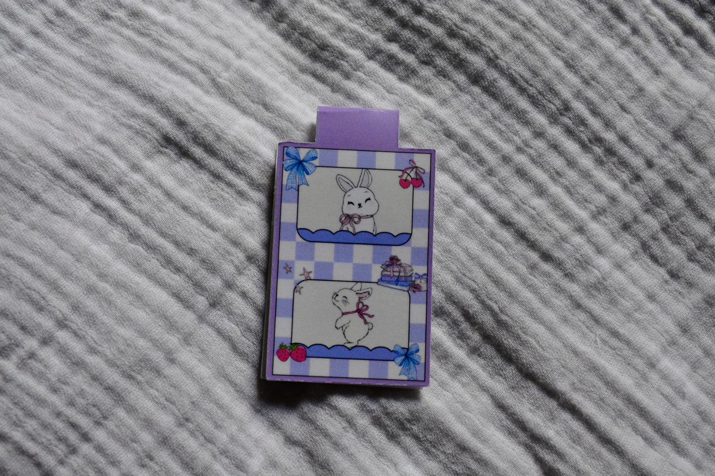 A small rectangular magnetic bookmark with a purple tab at the top, featuring a lavender-and-white gingham border and cute bunny illustrations, placed on a gray textured fabric background.
