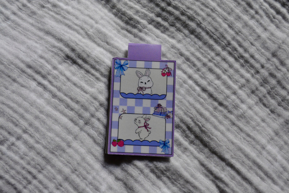 A small rectangular magnetic bookmark with a purple tab at the top, featuring a lavender-and-white gingham border and cute bunny illustrations, placed on a gray textured fabric background.