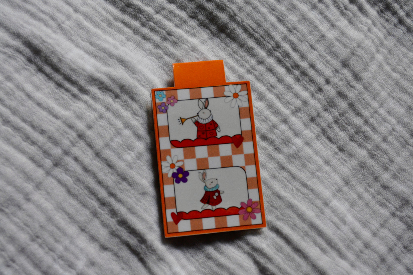 A small rectangular magnetic bookmark with an orange tab at the top, featuring a white bunny in a red outfit on an orange-and-white checkered background with floral accents, placed on a gray textured fabric surface.