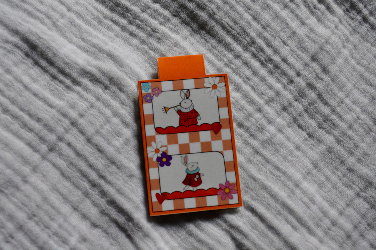 A small rectangular magnetic bookmark with an orange tab at the top, featuring a white bunny in a red outfit on an orange-and-white checkered background with floral accents, placed on a gray textured fabric surface.