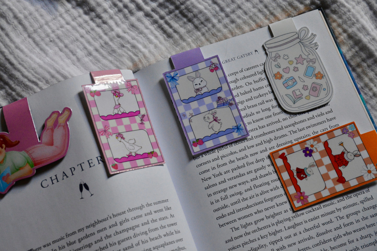 A set of colorful, whimsical magneticbookmarks styled on an open book. The bookmarks feature playful illustrations including a pink-and-white checkered bunny, a purple-and-white checkered cat, a gray bunny in a basket, a red-and-orange bunny with flowers, and a pink bunny with a bow.