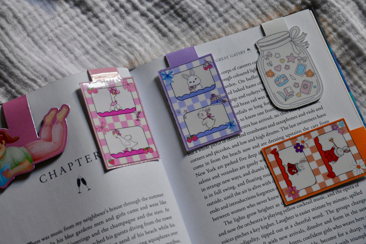 A set of colorful, whimsical magneticbookmarks styled on an open book. The bookmarks feature playful illustrations including a pink-and-white checkered bunny, a purple-and-white checkered cat, a gray bunny in a basket, a red-and-orange bunny with flowers, and a pink bunny with a bow.
