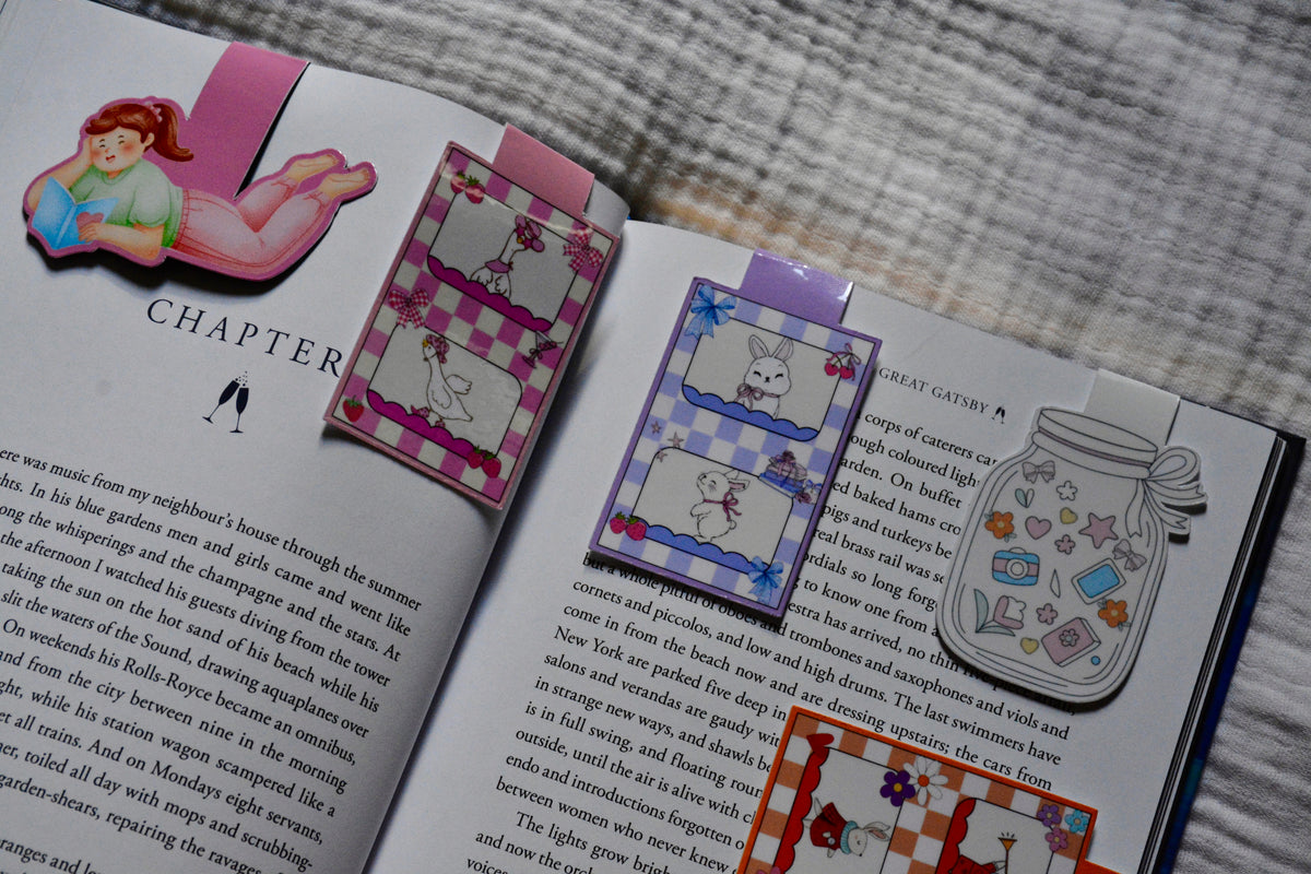 Three handmade magnetic bookmarks with illustrations of a girl, a bunnies, and a jar, displayed on open books.
