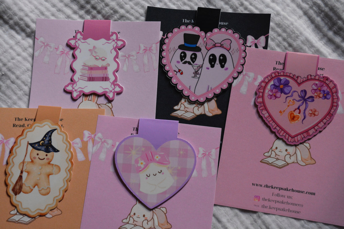 A set of cute, pastel-colored magnetic bookmarks or cards featuring cartoon characters, including a gingerbread bear in a witch hat, a ghost couple, and a bunny reading, arranged on a textured gray fabric background.