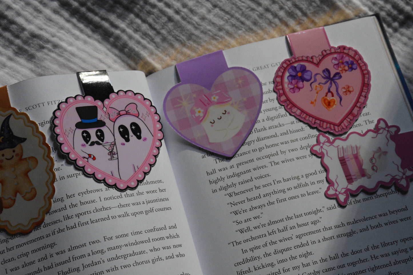 Three heart-shaped bookmarks with cartoon characters and illustrations are placed on open book pages. One bookmark is pink with a gingerbread wearing a top hat, one is purple with a cute ghost and one is pink with a books and sleeping ghost.