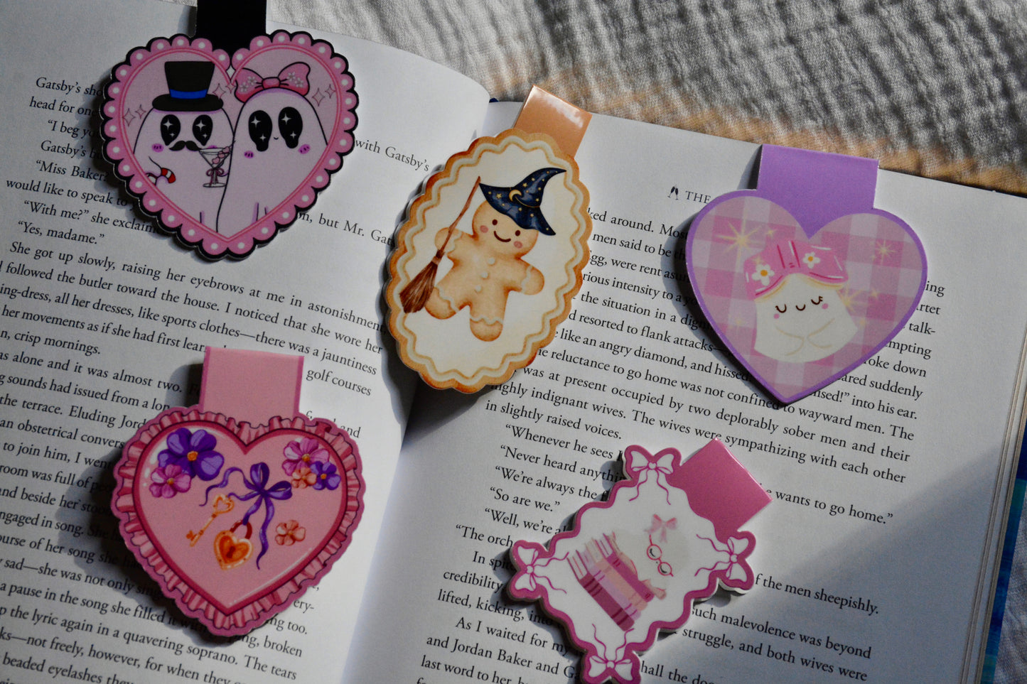 Three decorative heart-shaped and oval magnetic bookmarks with cartoon faces and pastel colors, placed on open book pages.