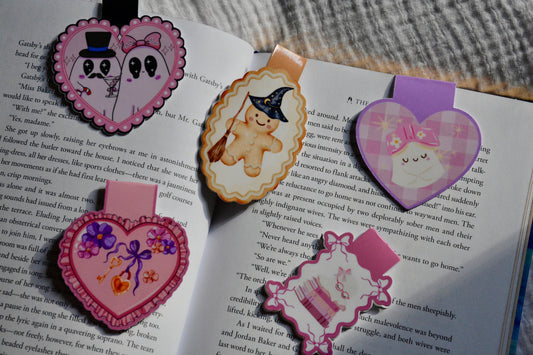 Three decorative heart-shaped and oval magnetic bookmarks with cartoon faces and pastel colors, placed on open book pages.