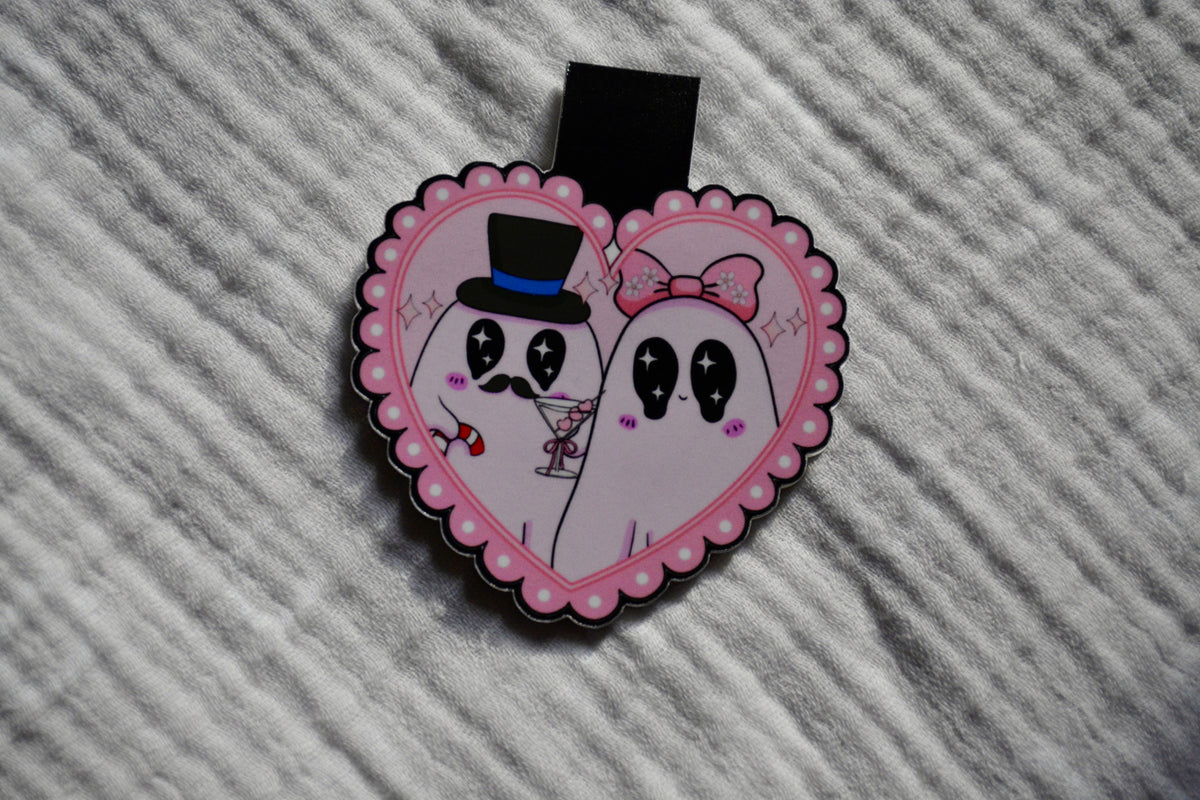 A heart-shaped enamel pin featuring two cute cartoon ghosts with black eyes, one wearing a black top hat and the other a pink bow, on a pink scalloped border, resting on a textured gray fabric background.