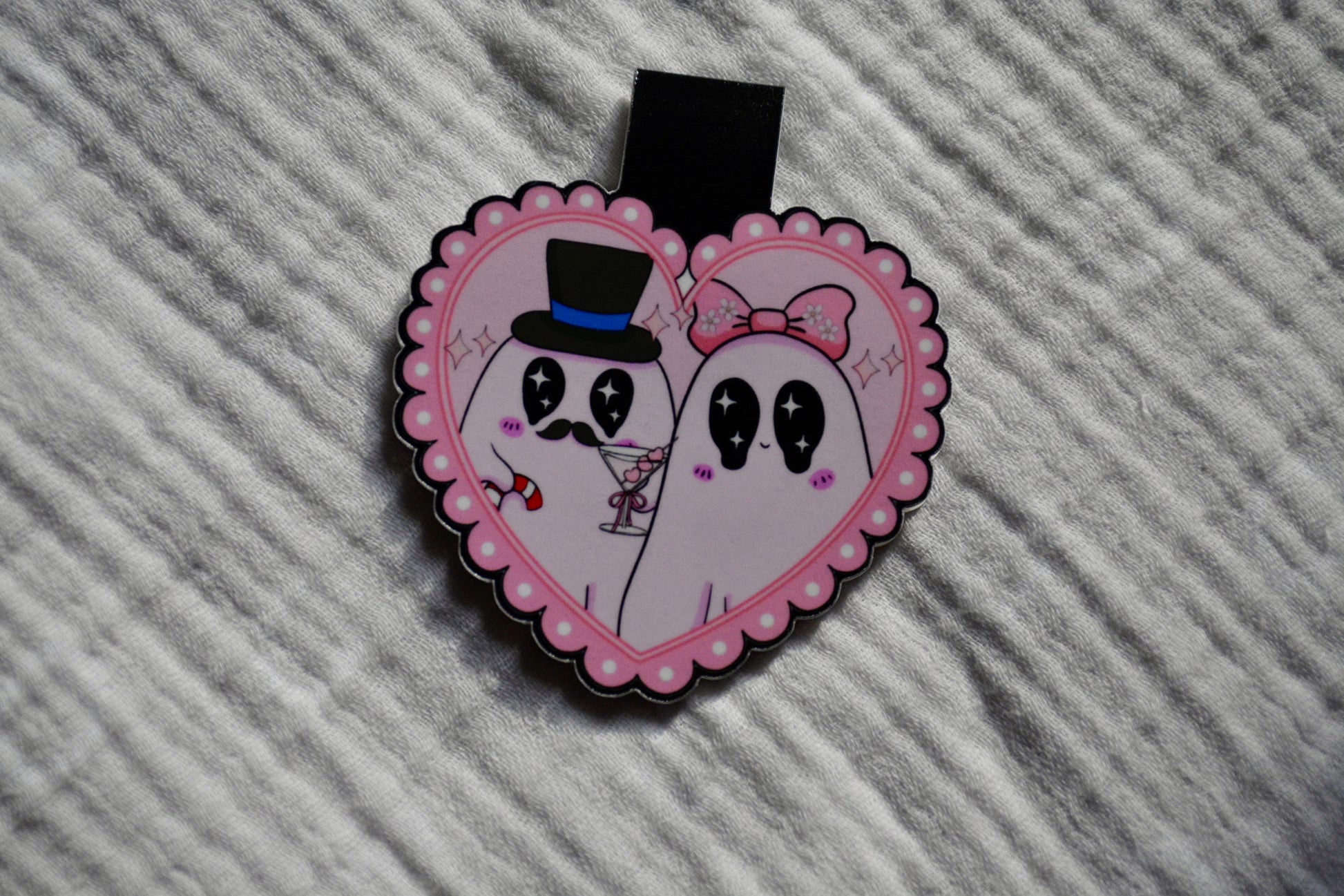 A heart-shaped enamel pin featuring two cute cartoon ghosts with black eyes, one wearing a black top hat and the other a pink bow, on a pink scalloped border, resting on a textured gray fabric background.