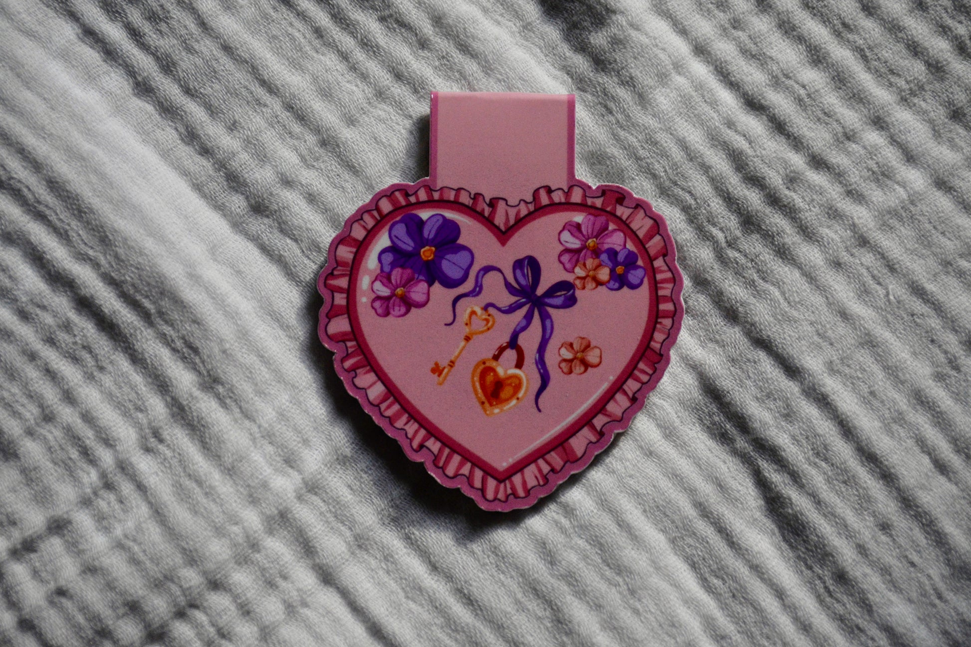 A heart-shaped magnetic bookmark with a decorative floral and ribbon illustration, placed on a textured gray fabric background.