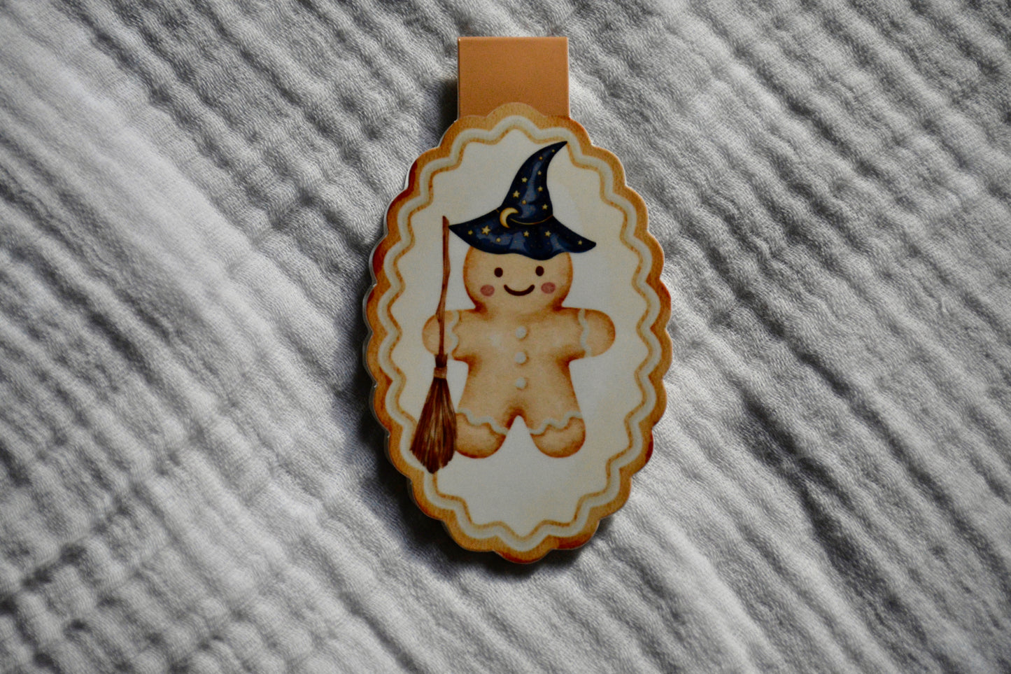 A Halloween-themed decorative magnetic featuring a gingerbread cookie wearing a witch hat and holding a broomstick, shown on a textured gray fabric background.