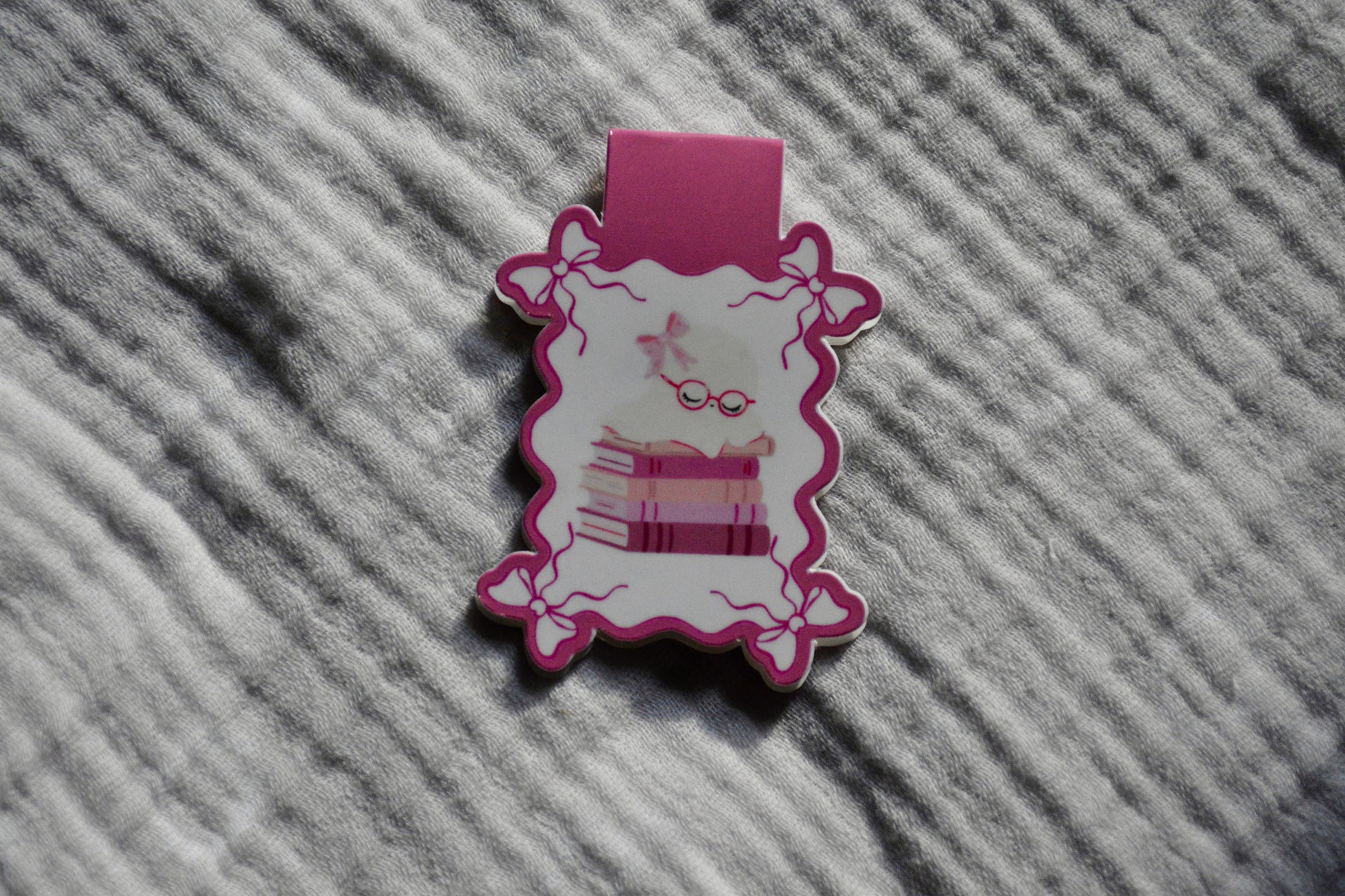 A small pink and white decorative accessory with a scalloped edge with stacked books and sleeping ghost, placed on a textured gray fabric background.