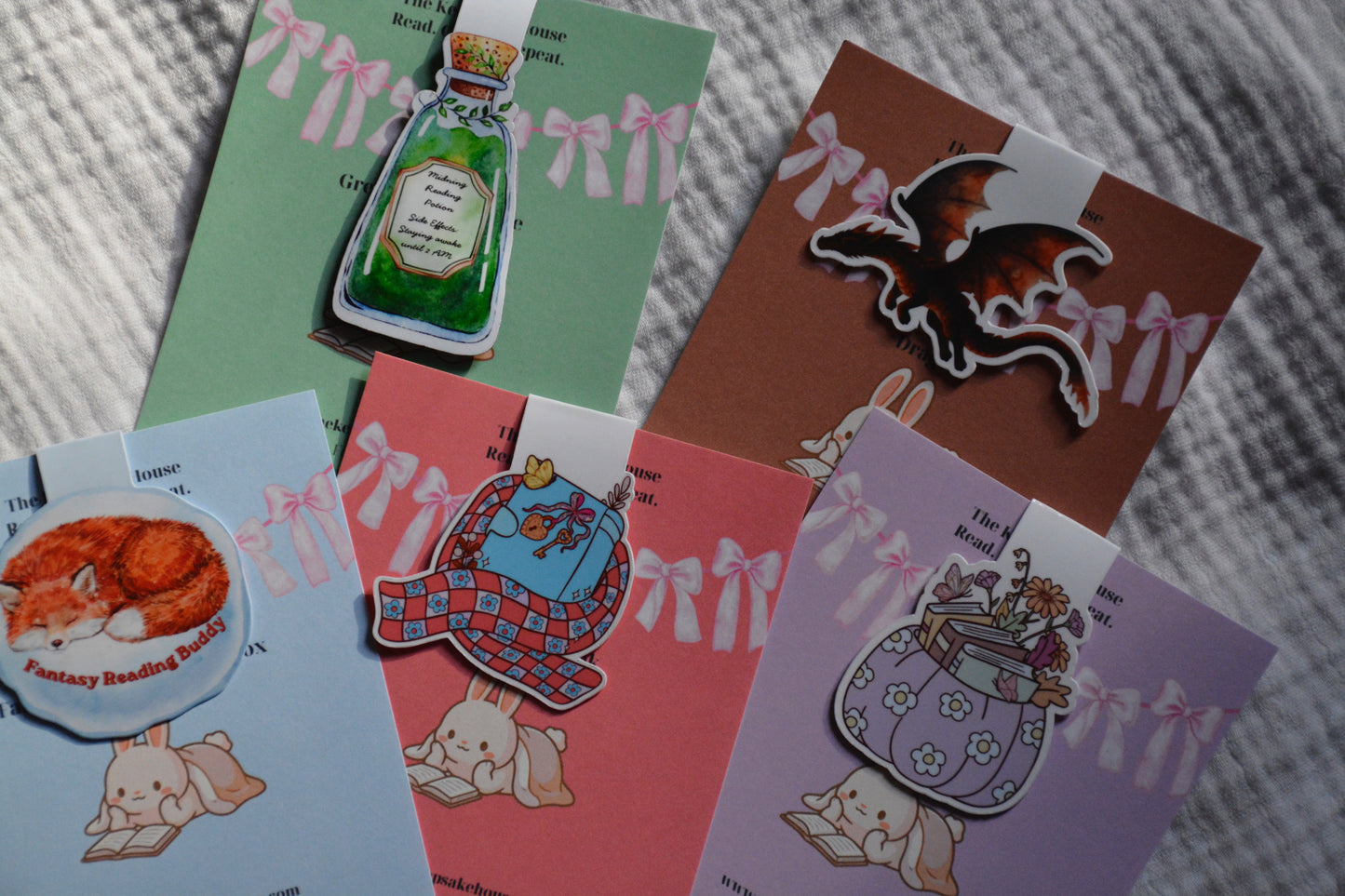 A collection of handmade magnetic bookmarks with cute illustrations, arranged on a gray textured blanket. The designs include a green potion bottle, a brown dragon, a blue bunny with a red plaid scarf, a purple bunny with a flower-patterned bag, and a light blue bunny with a cat sticker.