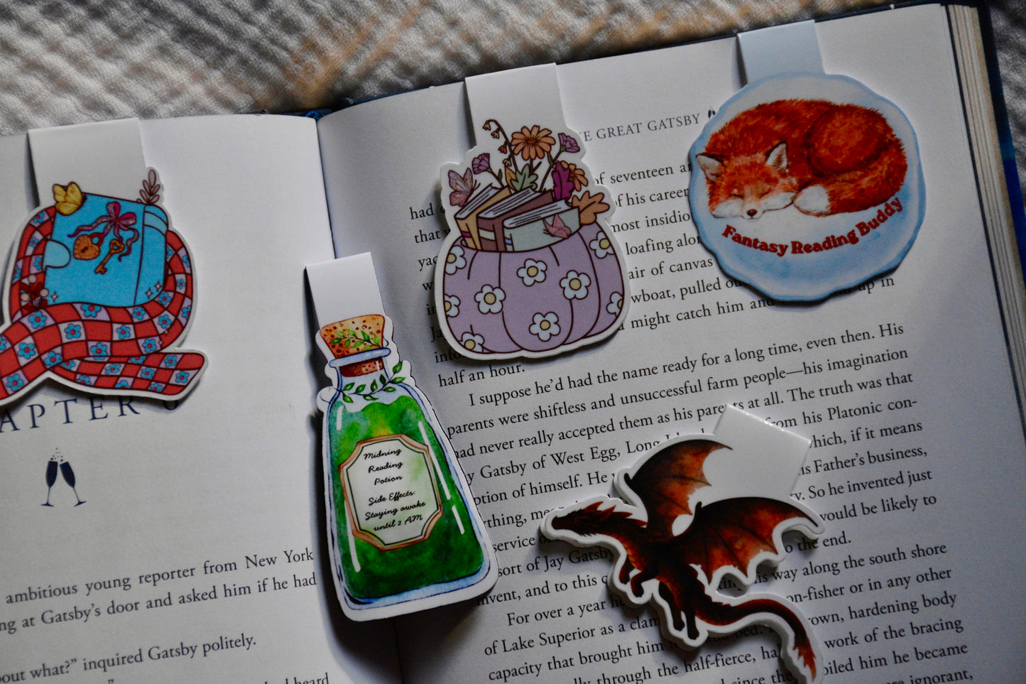 A set of fantasy inspired magnetic bookmarks styled on an open book. The bookmarks feature colorful illustrations including a red-and-blue plaid picnic blanket, a purple flower-patterned potion bottle, a blue fox head with 'Fantasy Reading Buddy' text, and a brown cat wearing a gold crown.