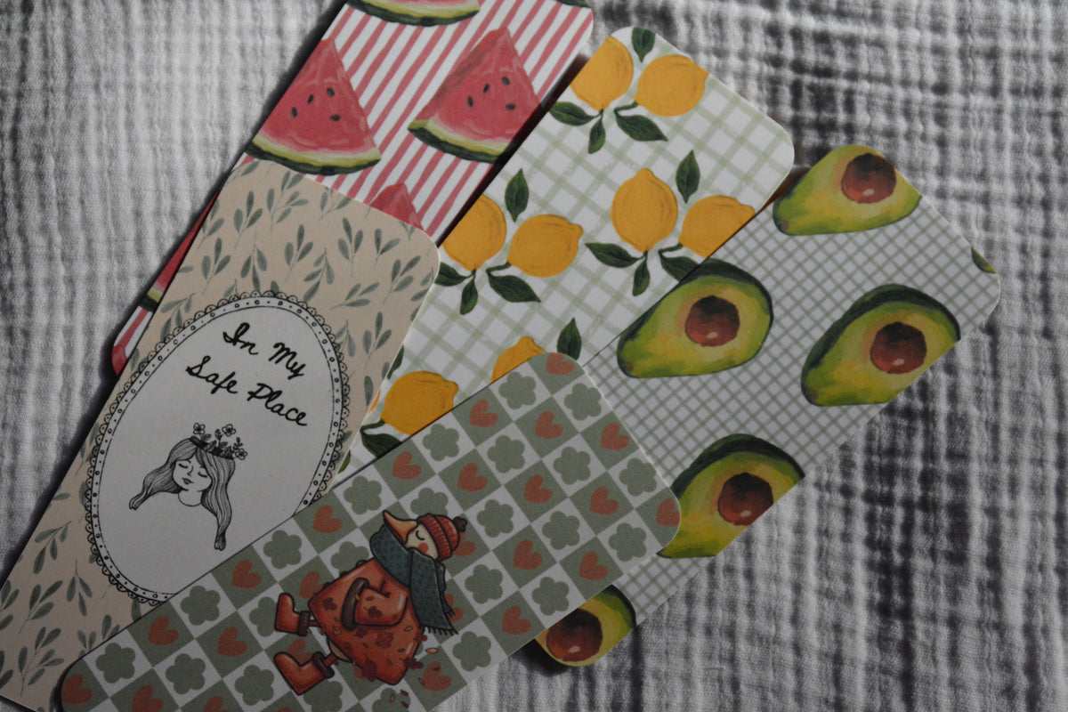 A set of patterned fabric bookmarks or tags fanned out on a gray textured fabric background. The designs include watermelons, lemons, avocados, a girl, and a cursive phrase 'In My Life Place' on a beige leafy background.