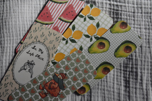A set of patterned fabric bookmarks or tags fanned out on a gray textured fabric background. The designs include watermelons, lemons, avocados, a girl, and a cursive phrase 'In My Life Place' on a beige leafy background.