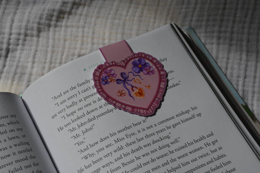 A heart-shaped pink magnetic bookmark with purple and orange floral designs and a bow, attached to an open book on a textured gray fabric background.