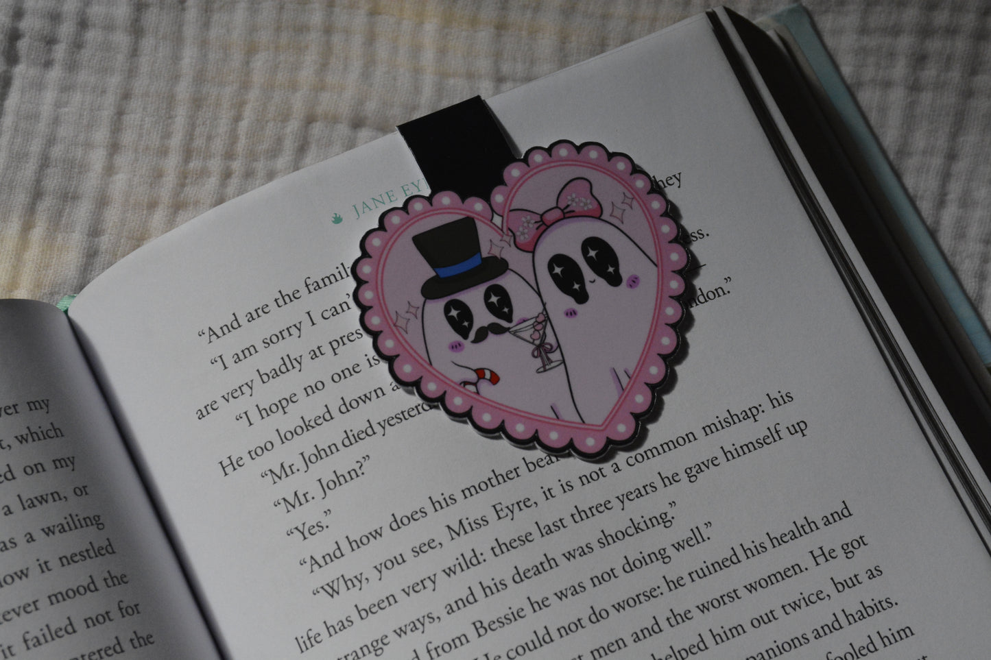 A heart-shaped pink  magnetic bookmark with a cartoon ghost couple illustration, placed on an open book page.