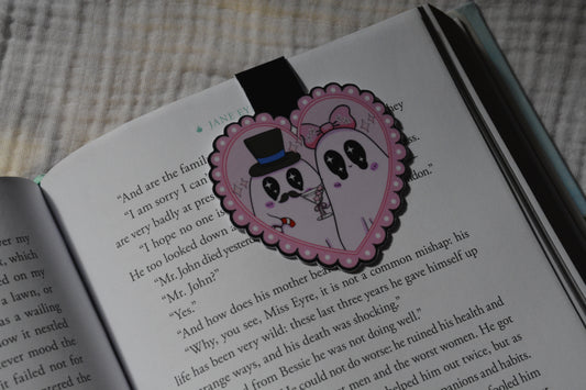 A heart-shaped pink  magnetic bookmark with a cartoon ghost couple illustration, placed on an open book page.