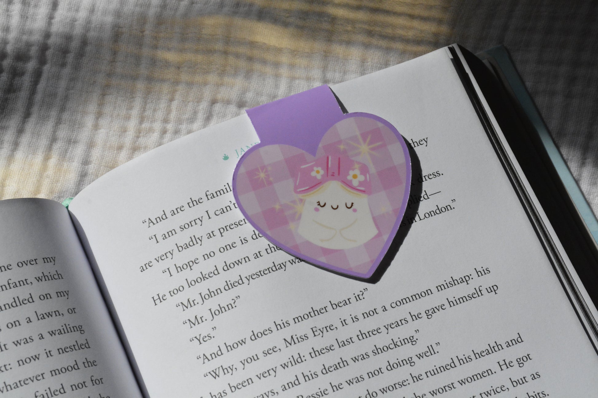 A purple heart-shaped bookmark with a plaid pattern and a cute sleeping character, placed on an open book.