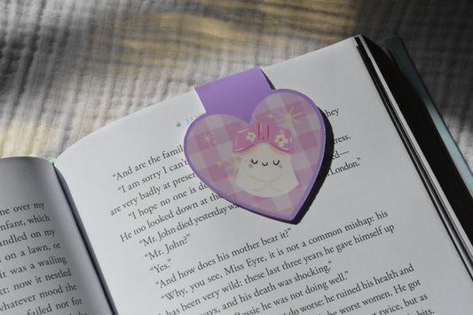 A purple heart-shaped bookmark with a plaid pattern and a cute sleeping character, placed on an open book.