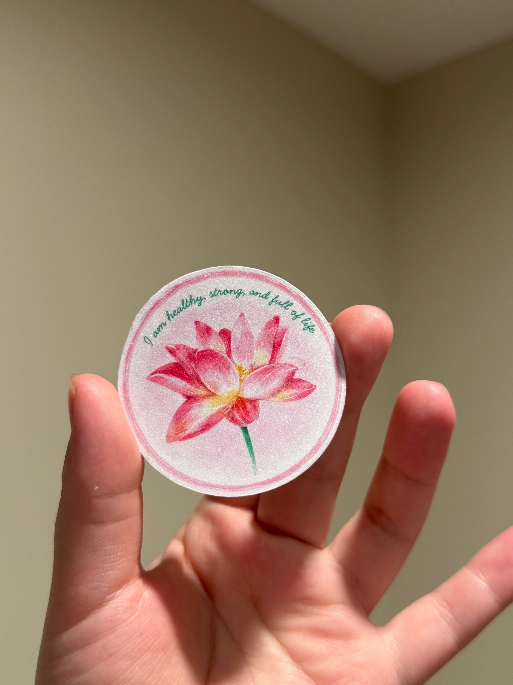 Handmade glitter affirmation sticker featuring a watercolor pink lotus flower with the quote "I am healthy, strong, and full of life," displayed in hand.