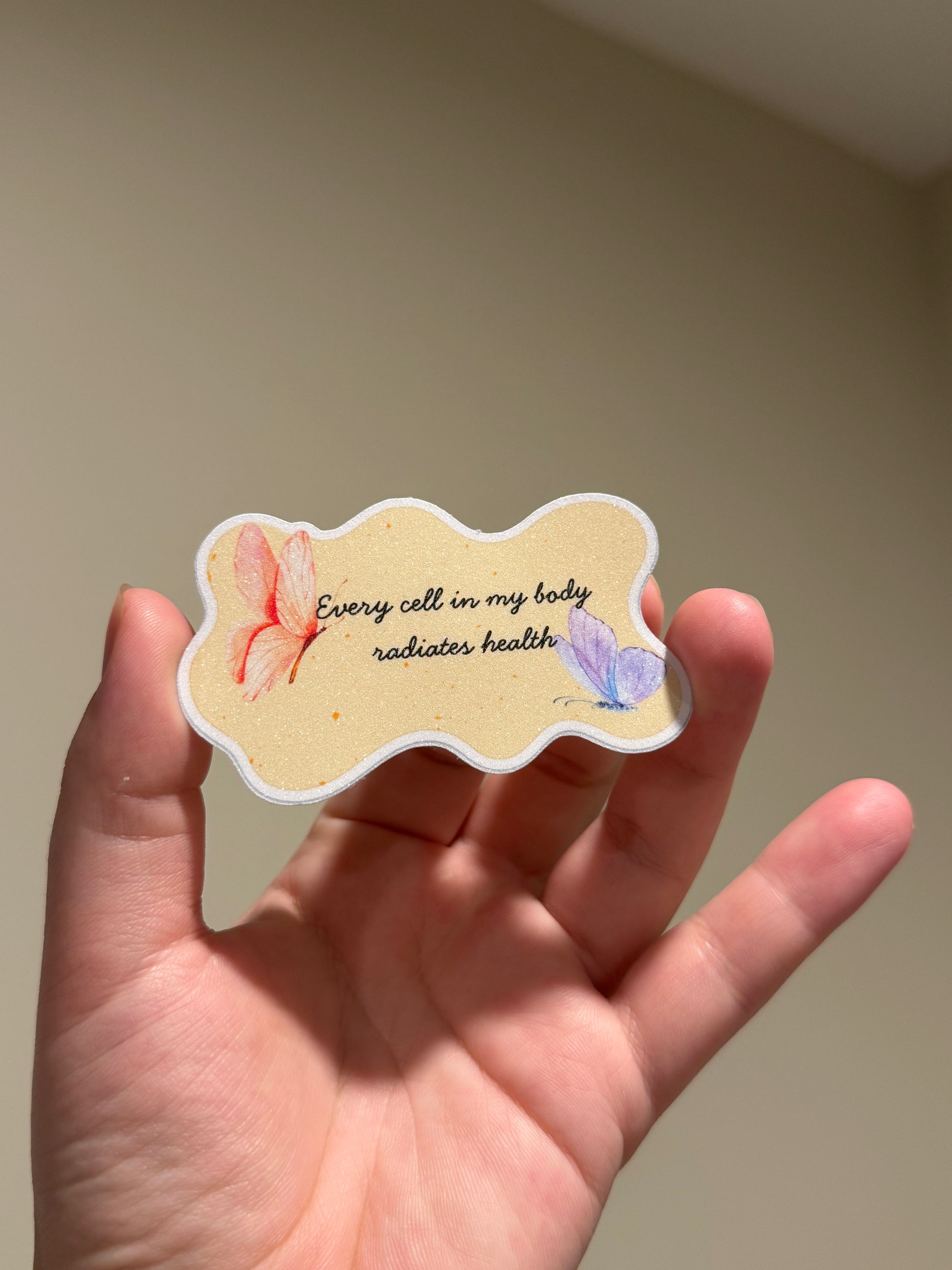 Handmade glitter affirmation sticker featuring watercolor butterflies and the quote "Every cell in my body radiates health," displayed in hand for Kindles, journals, laptops, and water bottles.