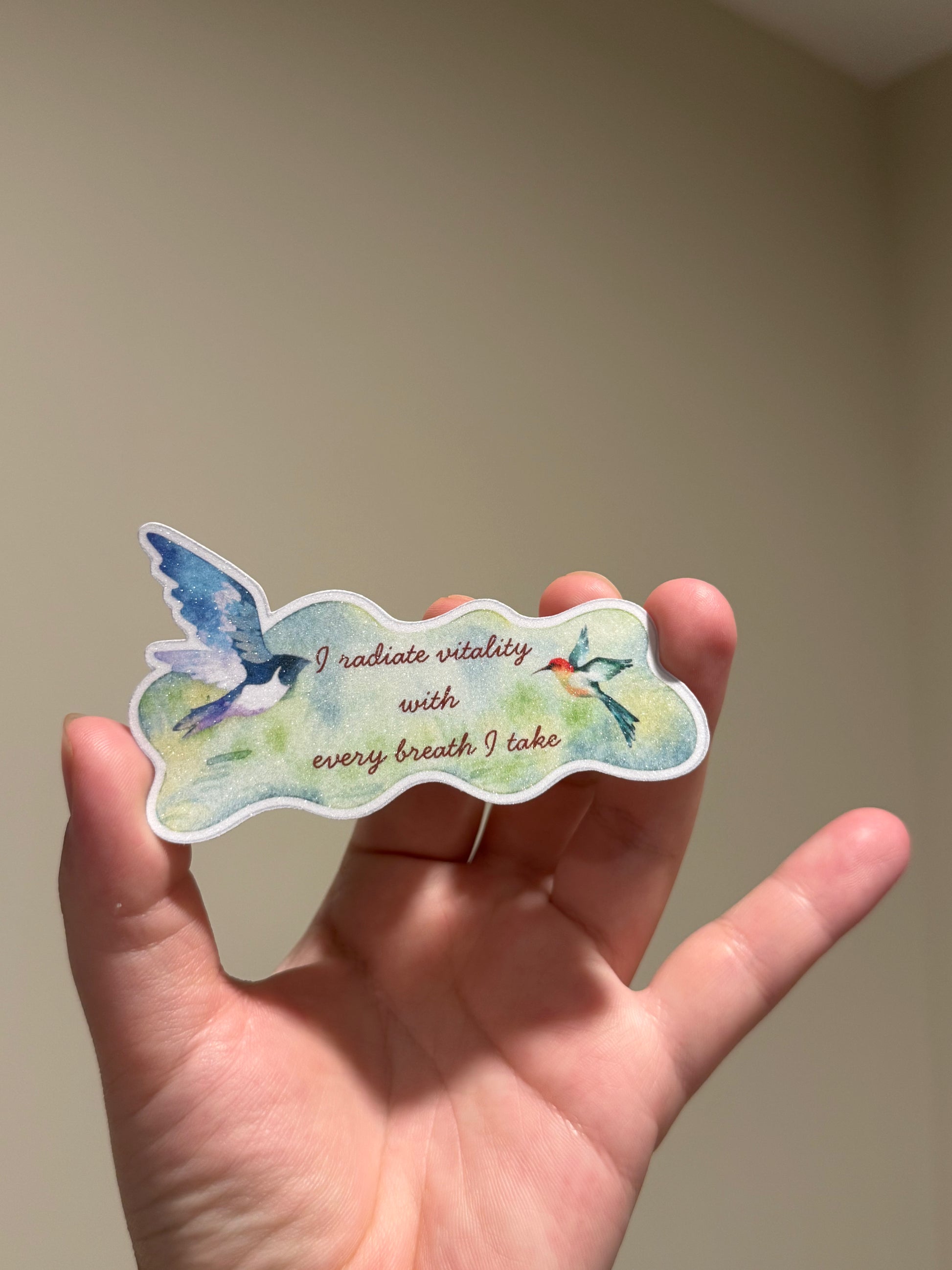 Handmade glitter affirmation sticker featuring watercolor birds and the quote "I radiate vitality with every breath I take," displayed in hand for Kindles, journals, laptops, and water bottles.