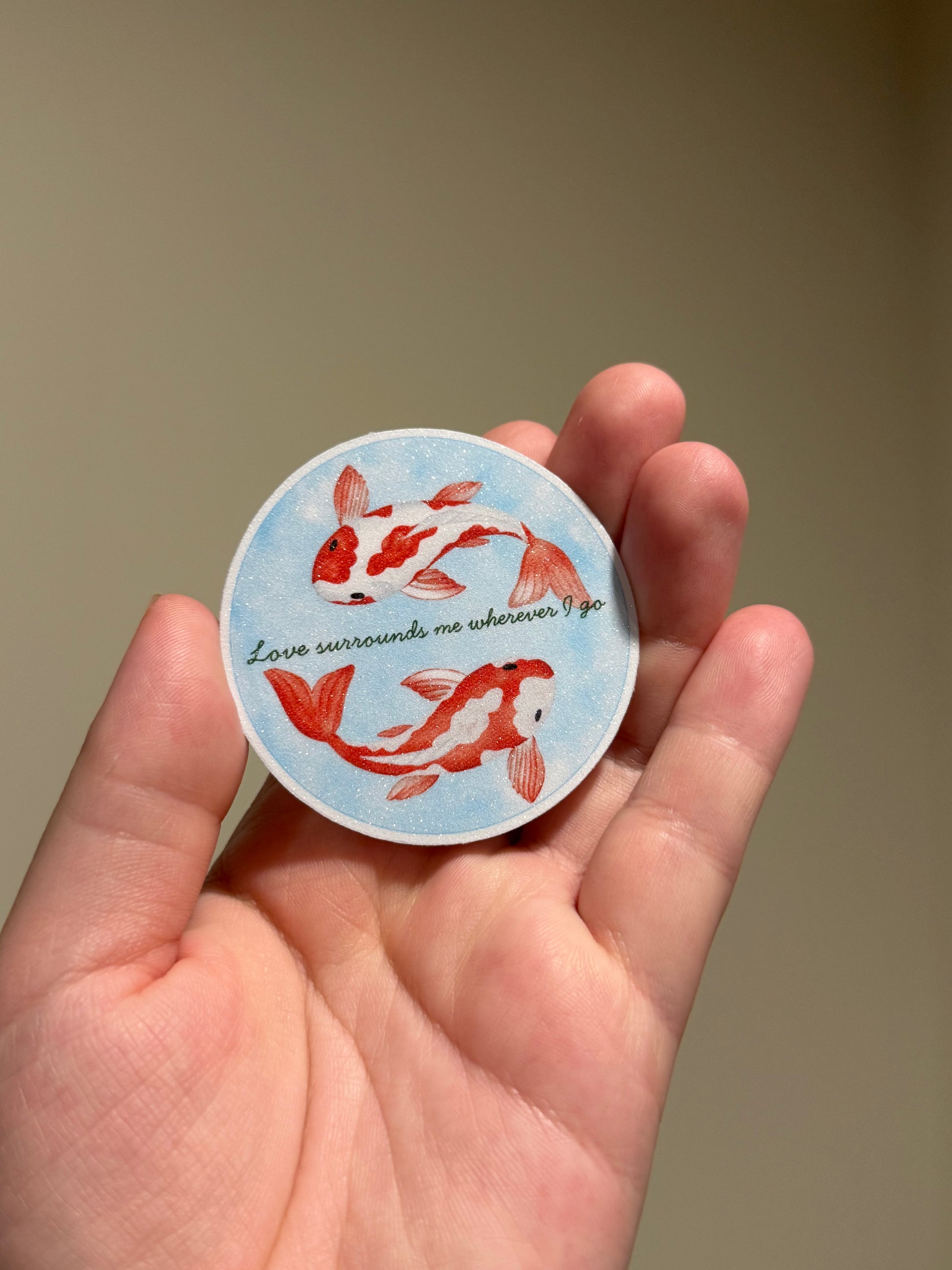Handmade glitter affirmation sticker featuring watercolor koi fish and the quote "Love surrounds me wherever I go," displayed in hand for Kindles, journals, laptops, and water bottles.