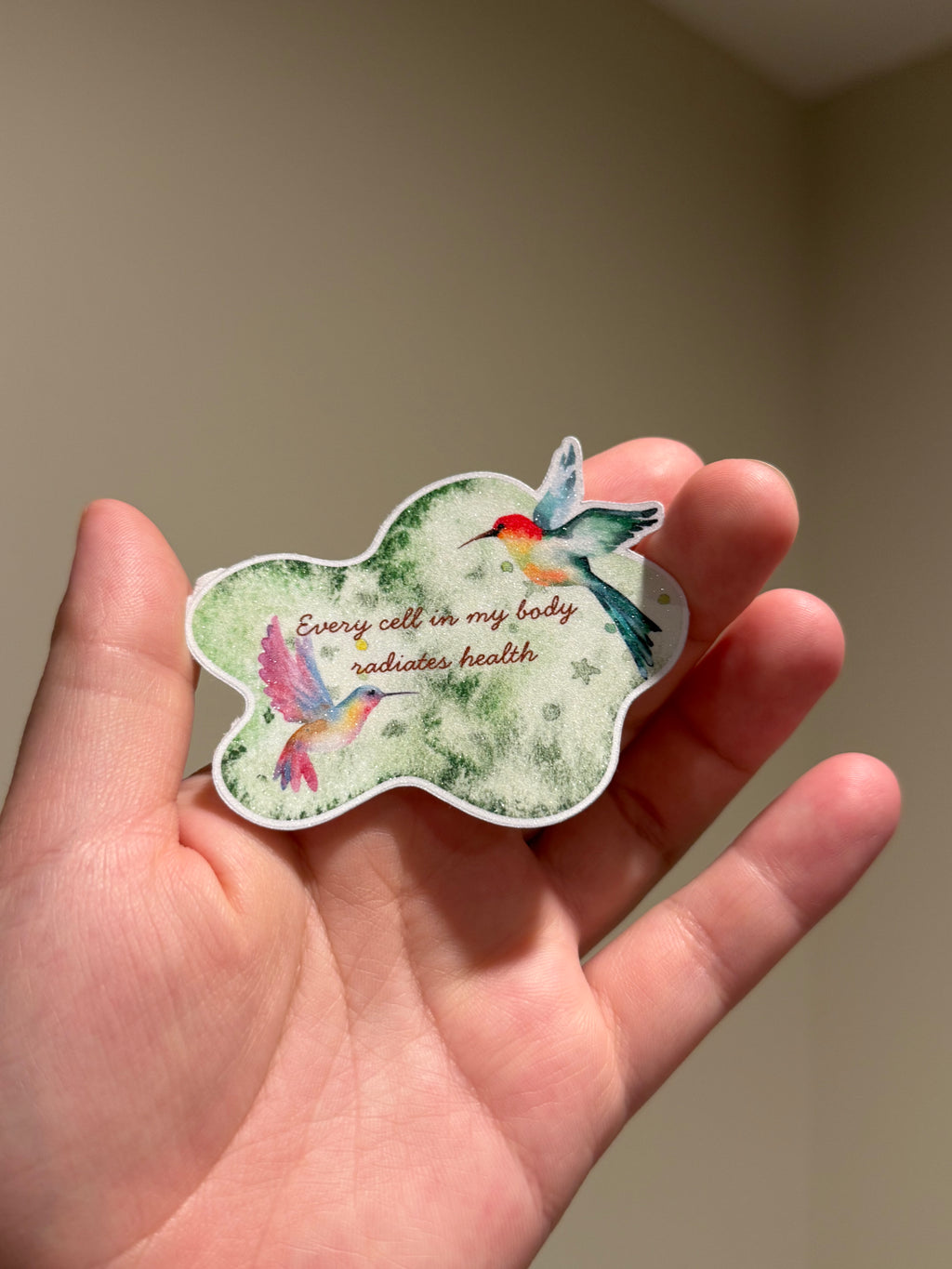 Handmade glitter affirmation sticker featuring watercolor hummingbirds and the quote "Every cell in my body radiates health," displayed in hand for Kindles, journals, laptops, and water bottles.