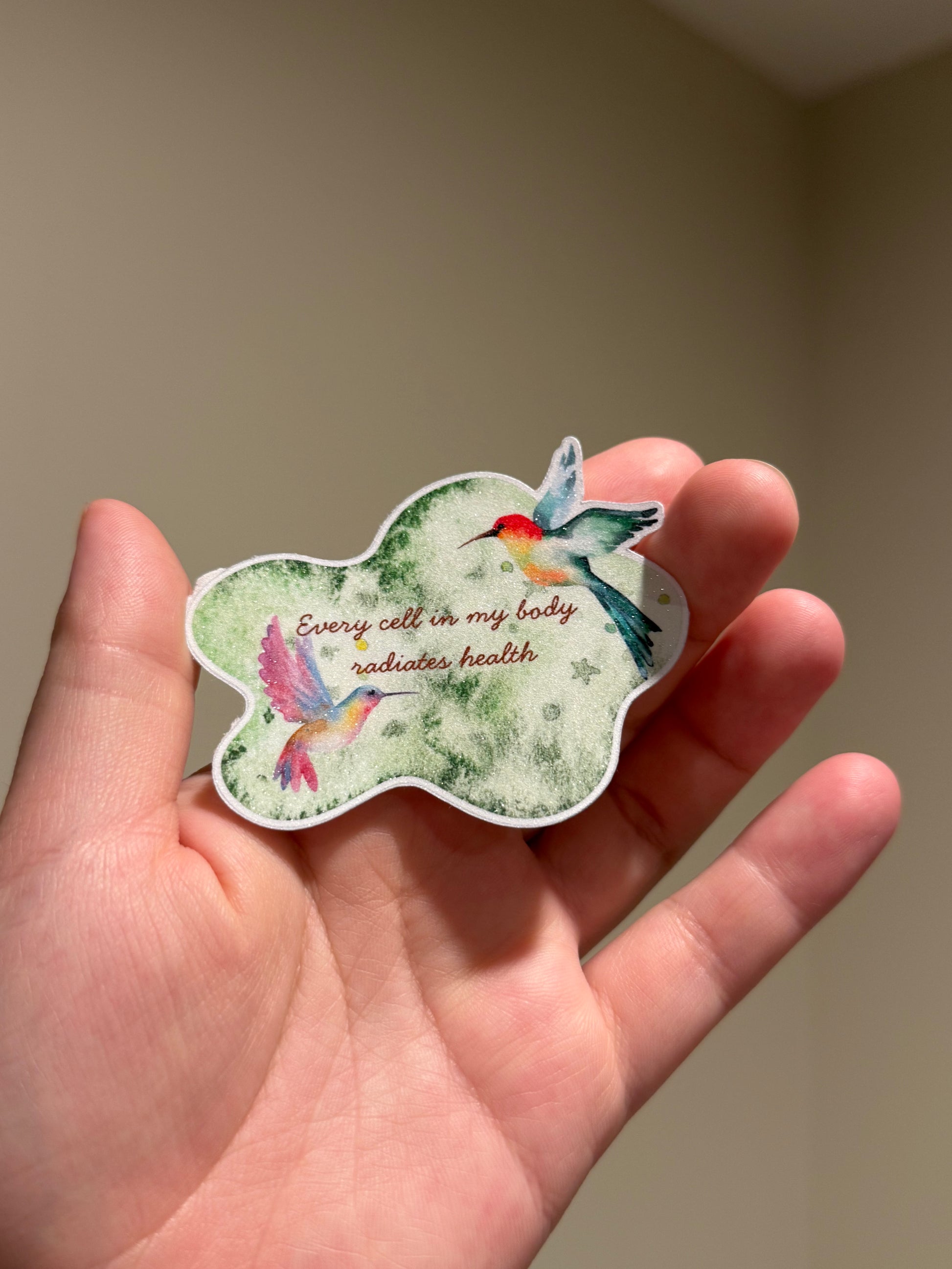 Handmade glitter affirmation sticker featuring watercolor hummingbirds and the quote "Every cell in my body radiates health," displayed in hand for Kindles, journals, laptops, and water bottles.