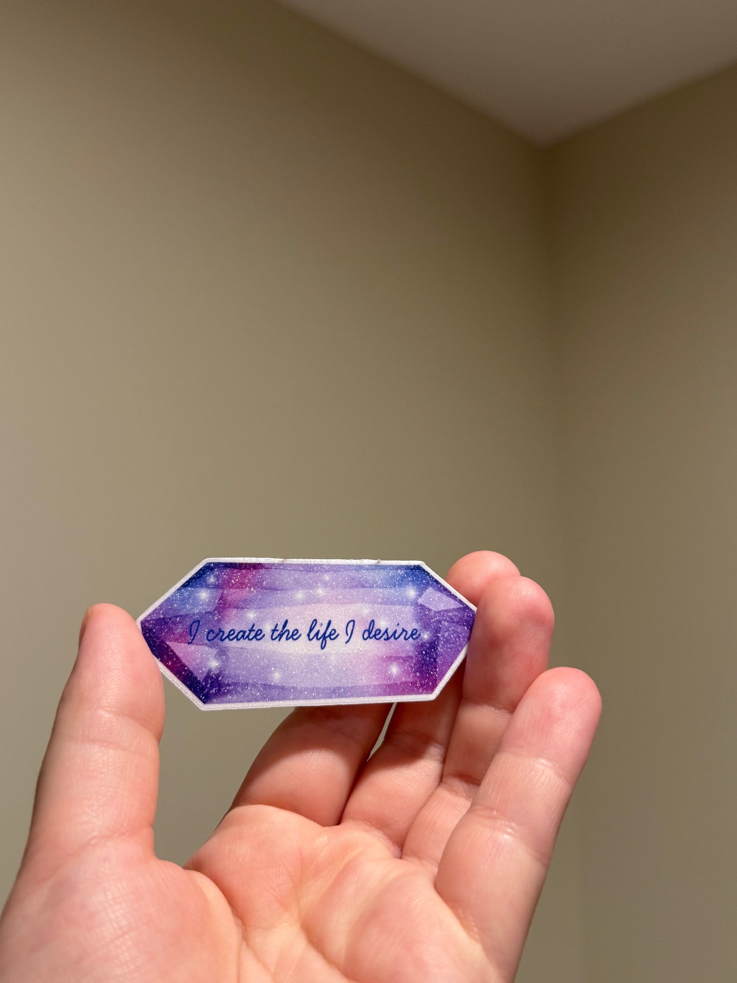 Purple glitter affirmation sticker featuring a crystal design and the quote "I create the life I desire," displayed in hand for Kindles, journals, planners, laptops, and water bottles.