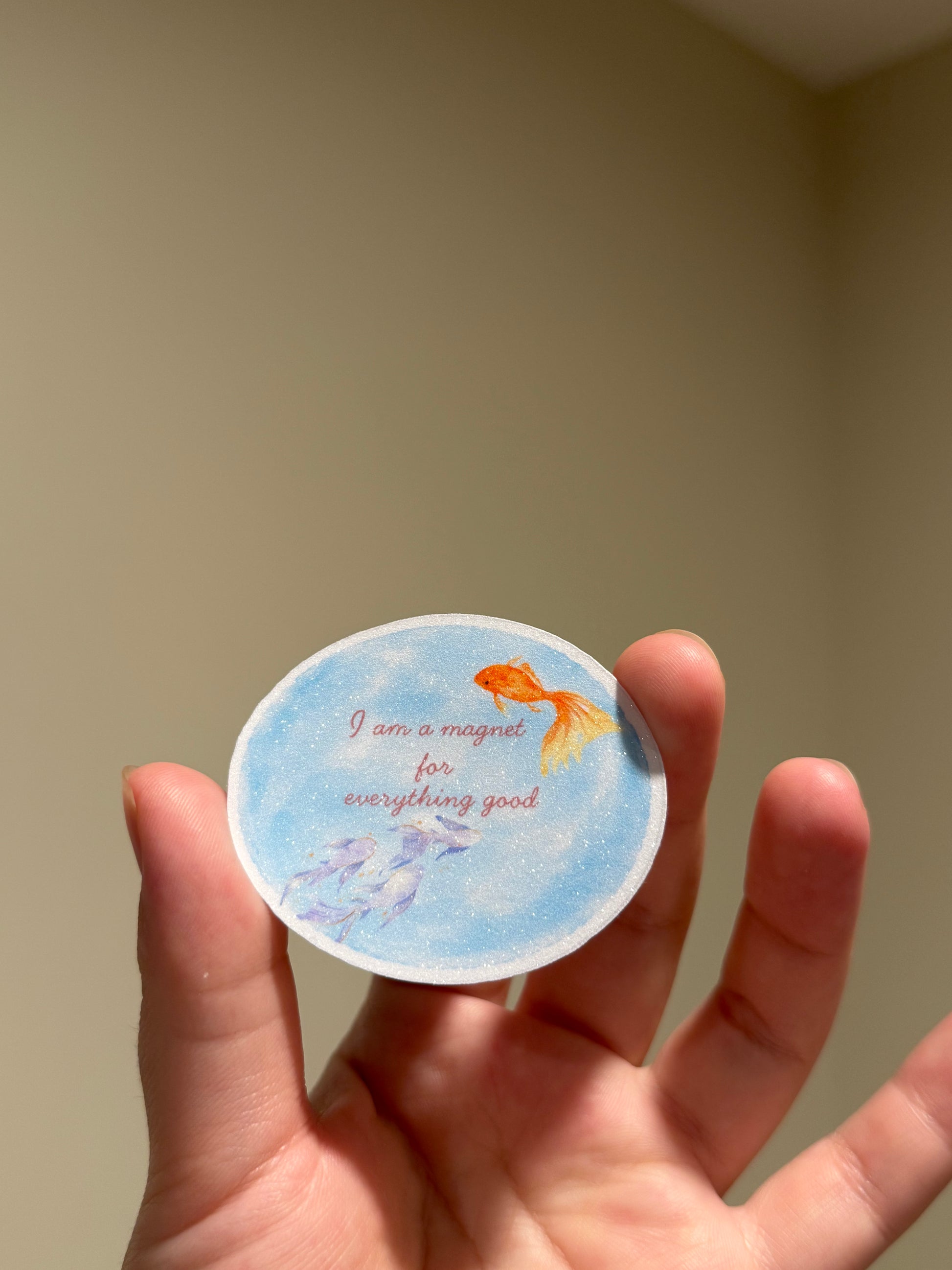 Blue glitter affirmation sticker featuring watercolor fish and the quote "I am a magnet for everything good" displayed in hand for Kindles, journals, planners, laptops, and water bottles.