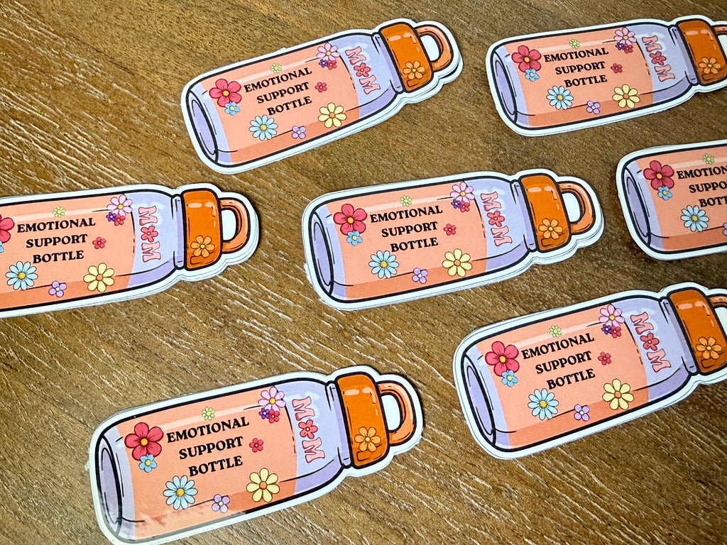 Several die-cut stickers shaped like illustrated pink bottles with orange caps and colorful flowers, printed with the text 'EMOTIONAL SUPPORT BOTTLE,' arranged on a wood surface.