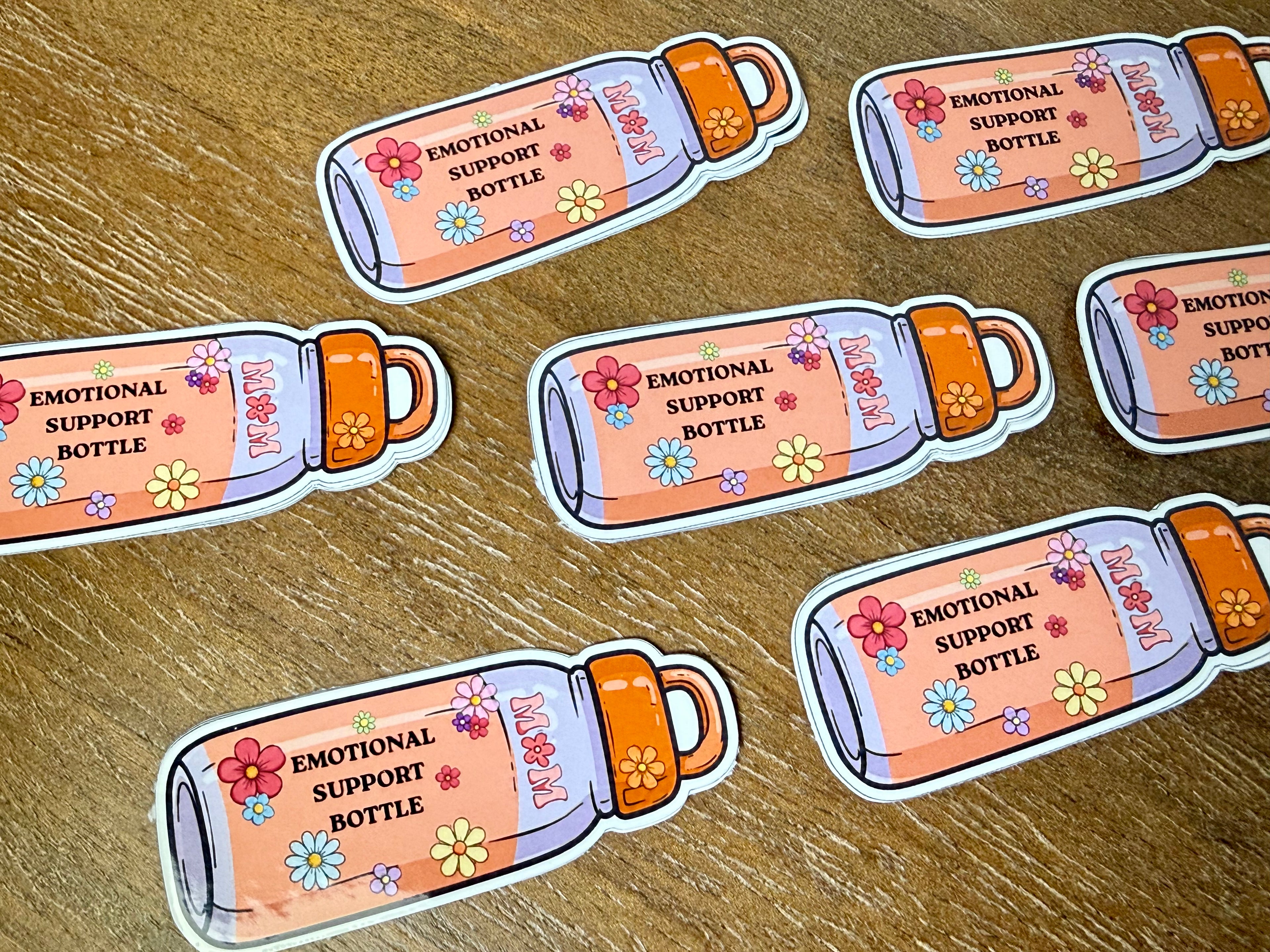 Several die-cut stickers shaped like illustrated pink bottles with orange caps and colorful flowers, printed with the text 'EMOTIONAL SUPPORT BOTTLE,' arranged on a wood surface.