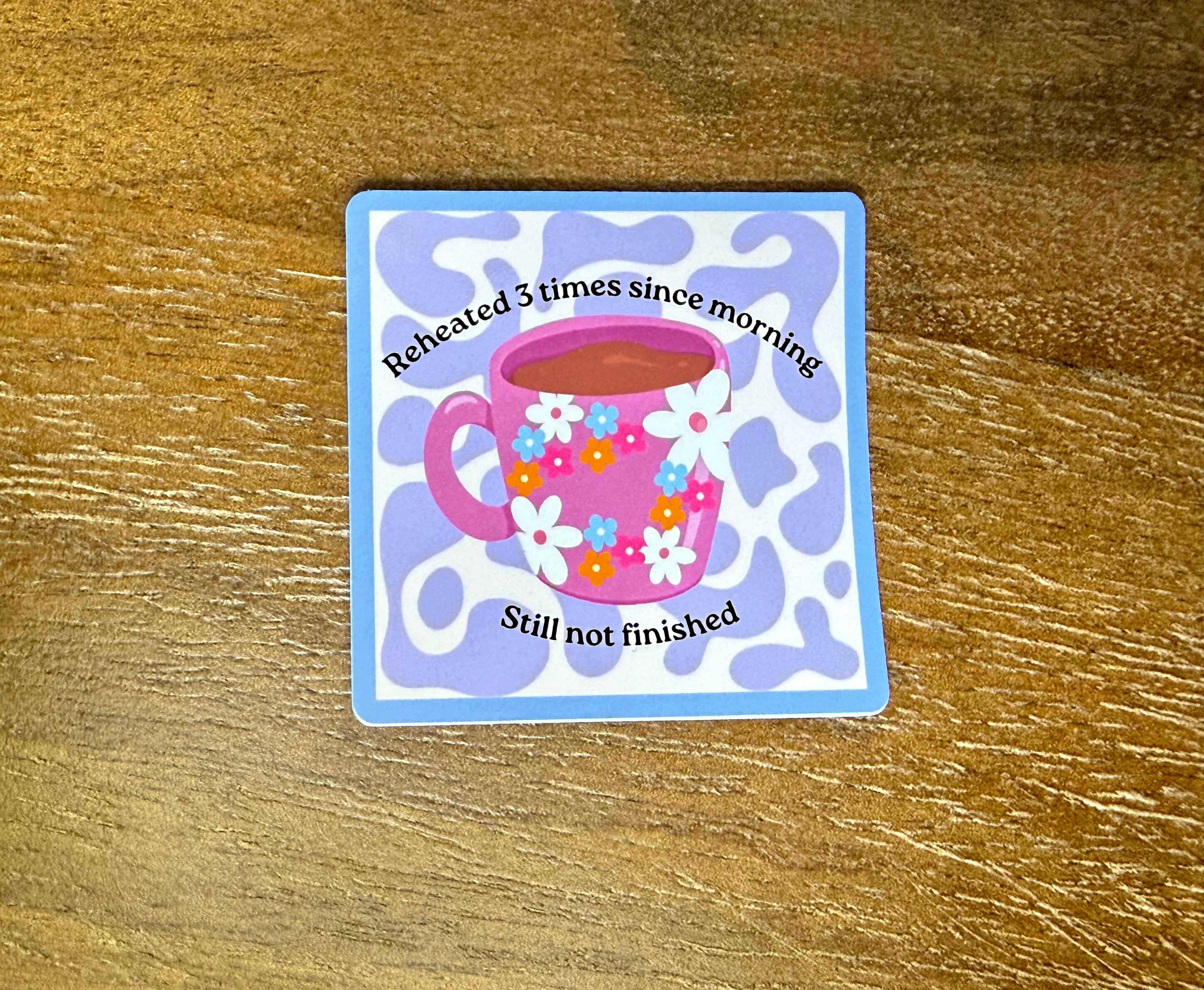 Funny "Reheated 3 Times Since Morning, Still Not Finished" coffee mug sticker with a floral mug illustration on a pastel purple background.