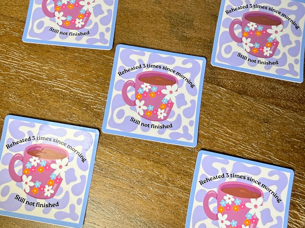 Five square printed stikers with rounded corners arranged on a wooden surface. Each card shows a pink floral teacup illustration and the text 'Reheated 3 times since morning' and 'Still not finished.