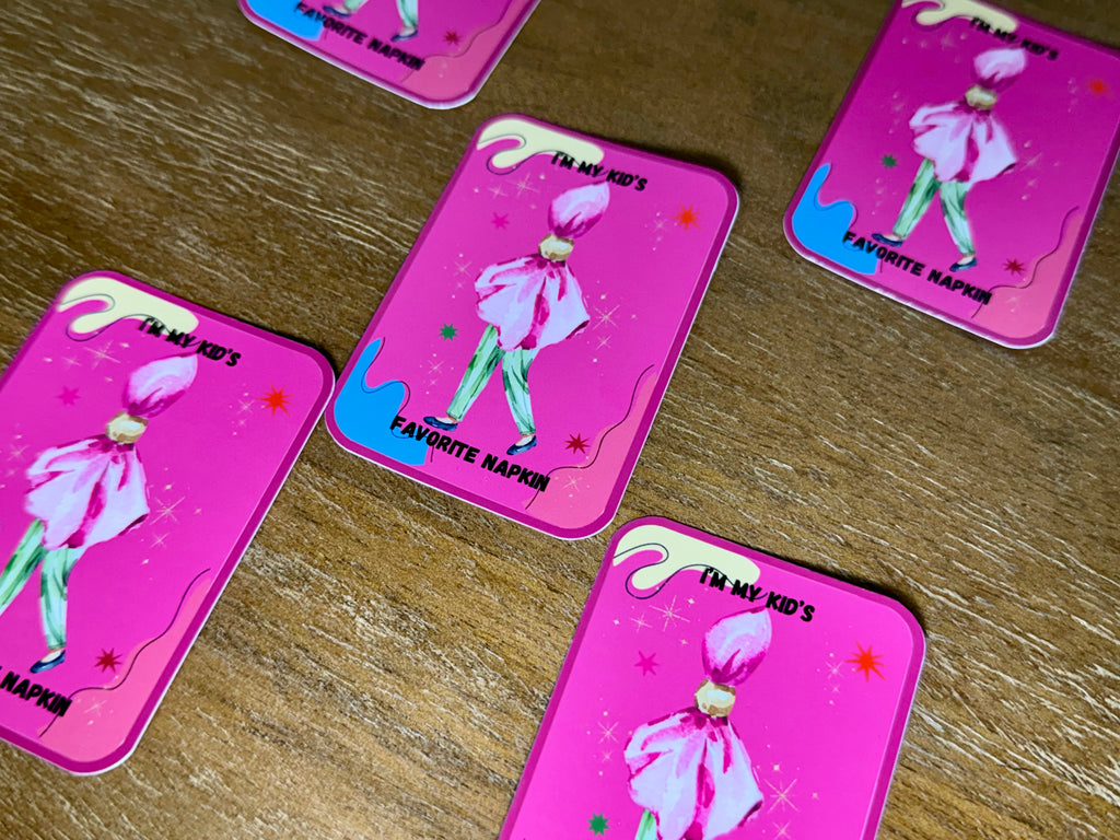 Pink rectangular cards with rounded corners arranged on a wooden surface. Each card shows a stylized figure in a pink bow and green pants with the text 'I'M MY KID'S' and 'FAVORITE NAPKIN'.